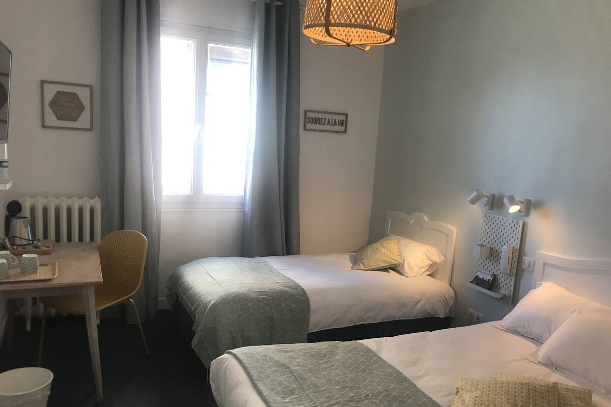 A hotel room with two beds, a desk with a chair, a radiator, and a window with curtains.