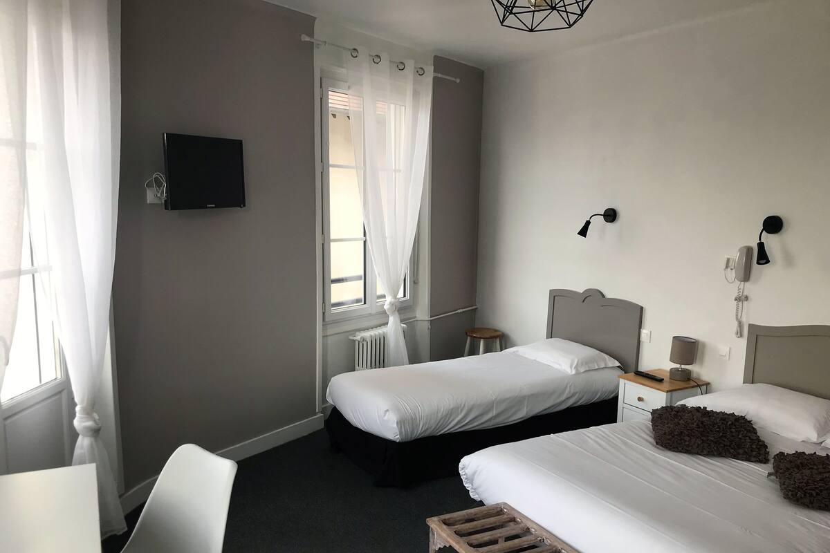 A hotel room with two beds, a TV, a desk, a chair, a small stool, and a window with curtains.