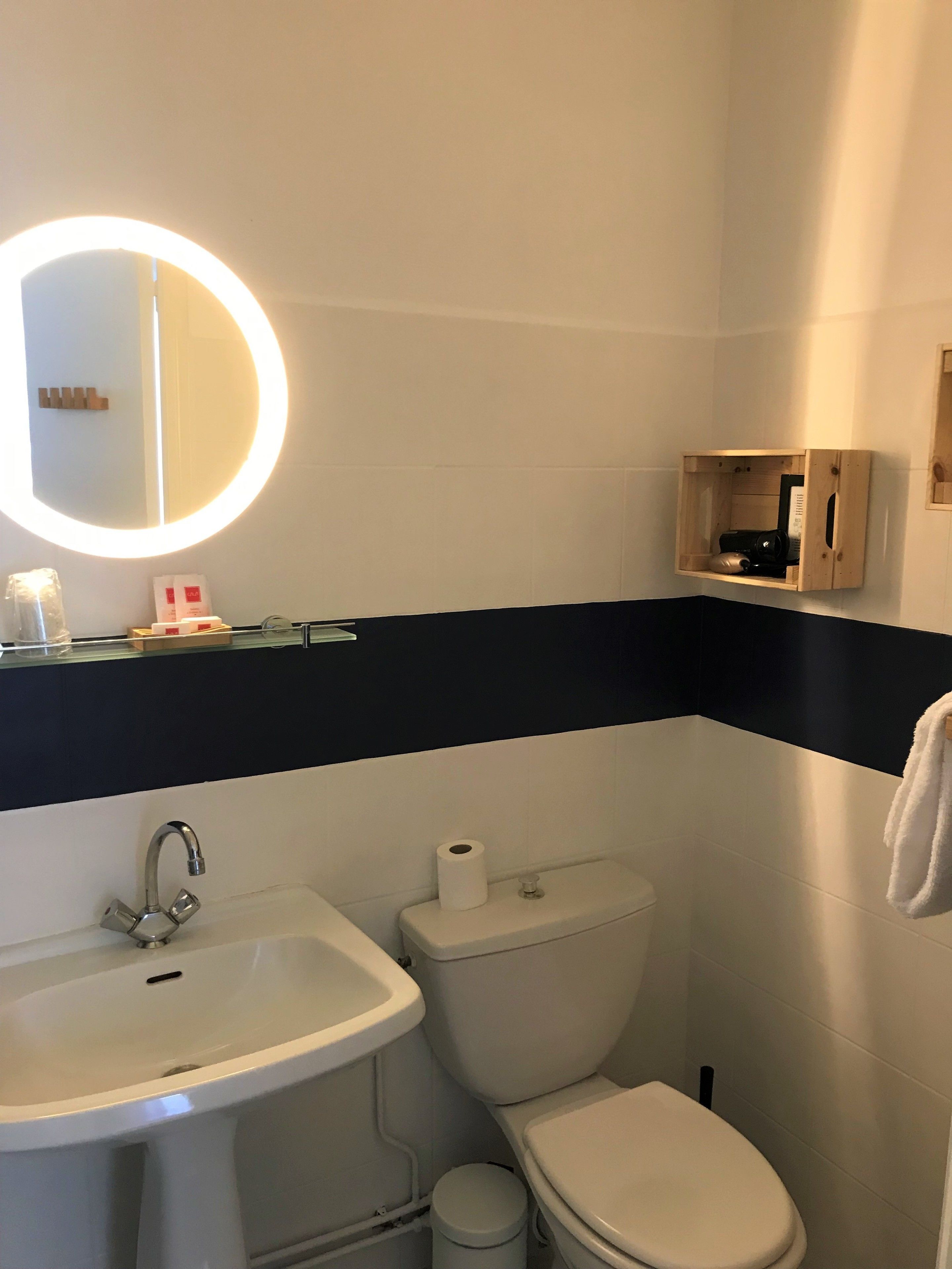 A bathroom with a sink, toilet, and a mirror with a light. There is a small wooden cabinet with toiletries.