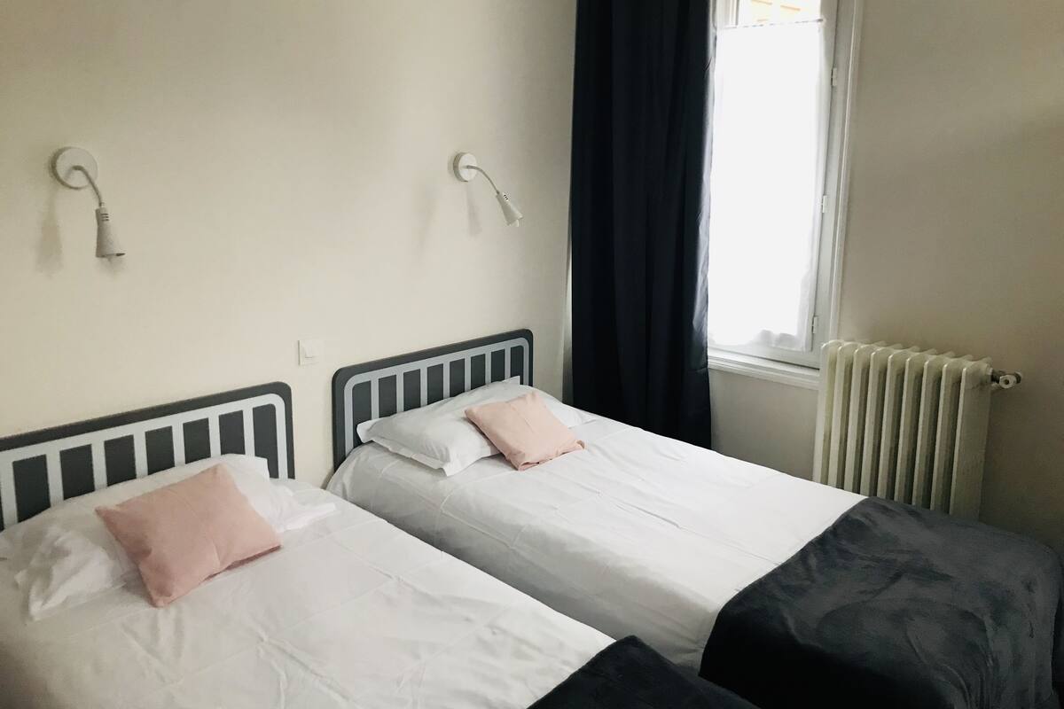 A bedroom with two beds, a window with curtains, a radiator, and a wall-mounted light fixture.