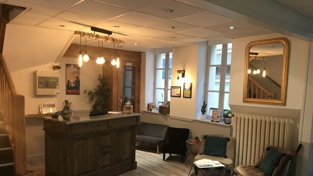 A spacious living area with a wooden floor, a dark wooden bar, and a large mirror. There are multiple seating options, including a sofa and two armchairs. - Hôtel L'ESTRAN (Trouville-sur-Mer)