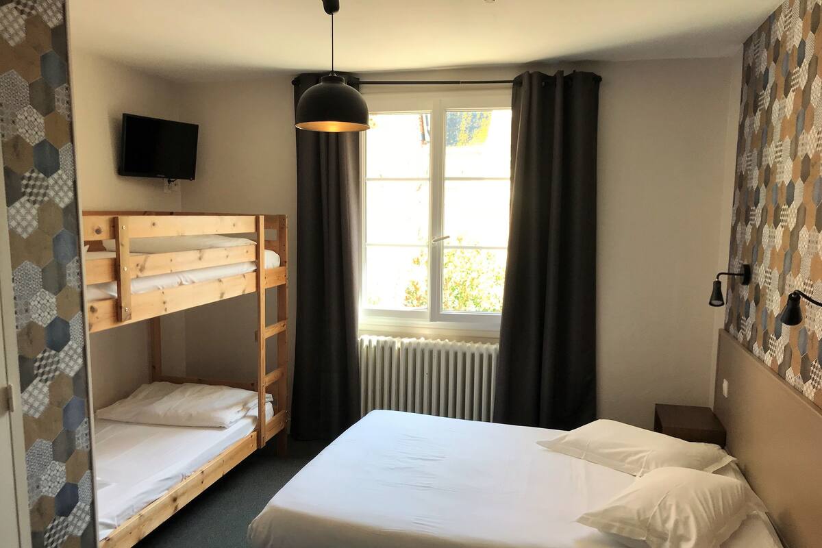 A room with a bunk bed, a single bed, a window with curtains, a radiator, and a wall-mounted TV.