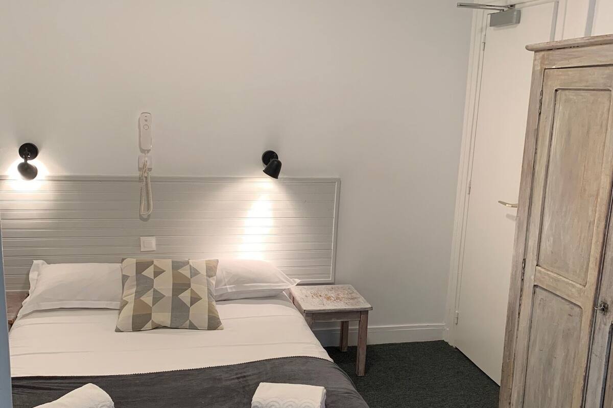 A neatly made bed with a dark grey bedspread and two white towels, a bedside table with a phone, and a wooden wardrobe in a simple room with white walls.