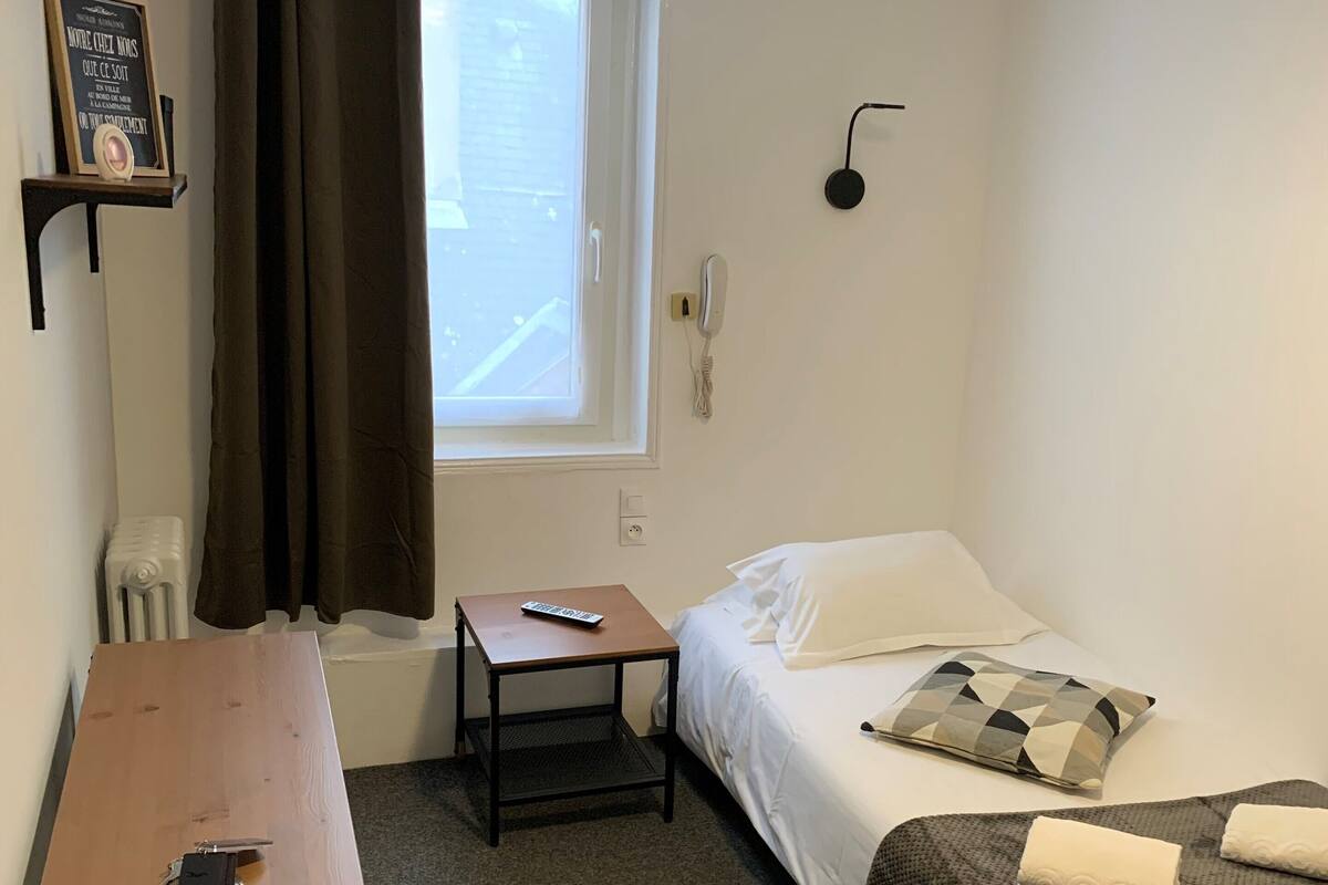 A small, neatly arranged room with a bed, a desk, and a chair. There is a window with curtains, a wall-mounted phone, and a shelf with a framed item.