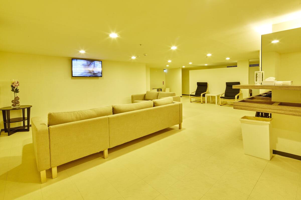 A modern hotel room with a beige sofa, a TV mounted on the wall, and a small table with a vase.