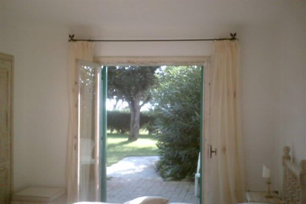 Une chambre à coucher avec un lit, une table de chevet et une vue sur un jardin, visible à travers une porte vitrée.