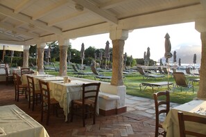 An outdoor restaurant with tables and chairs, covered by a roof, and overlooking a pool area. - IHR Hotel San Paolo (Loiri Porto San Paolo)