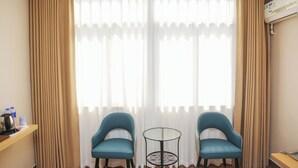 A hotel room with two blue chairs, a small round table, a minibar, and a large window with sheer curtains. - Green Oasis Hotel (Shenzhen)