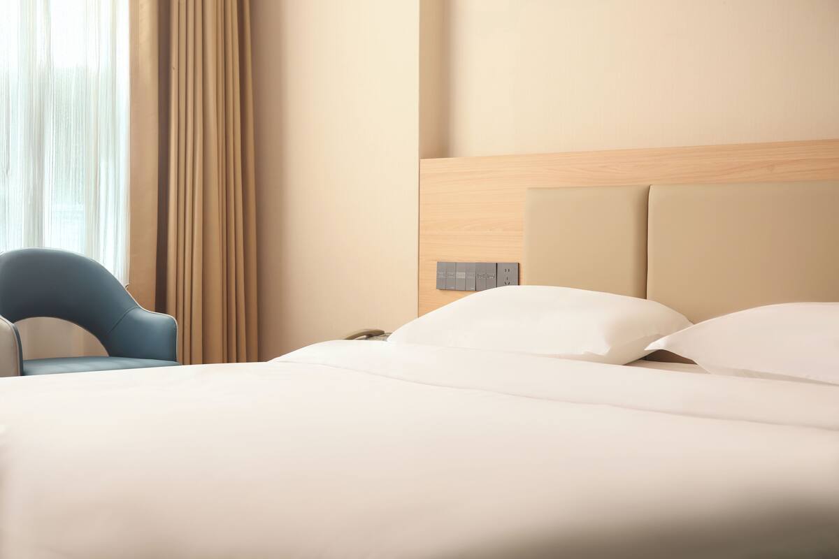A hotel room with a bed, a chair, a nightstand, and a wall-mounted control panel.
