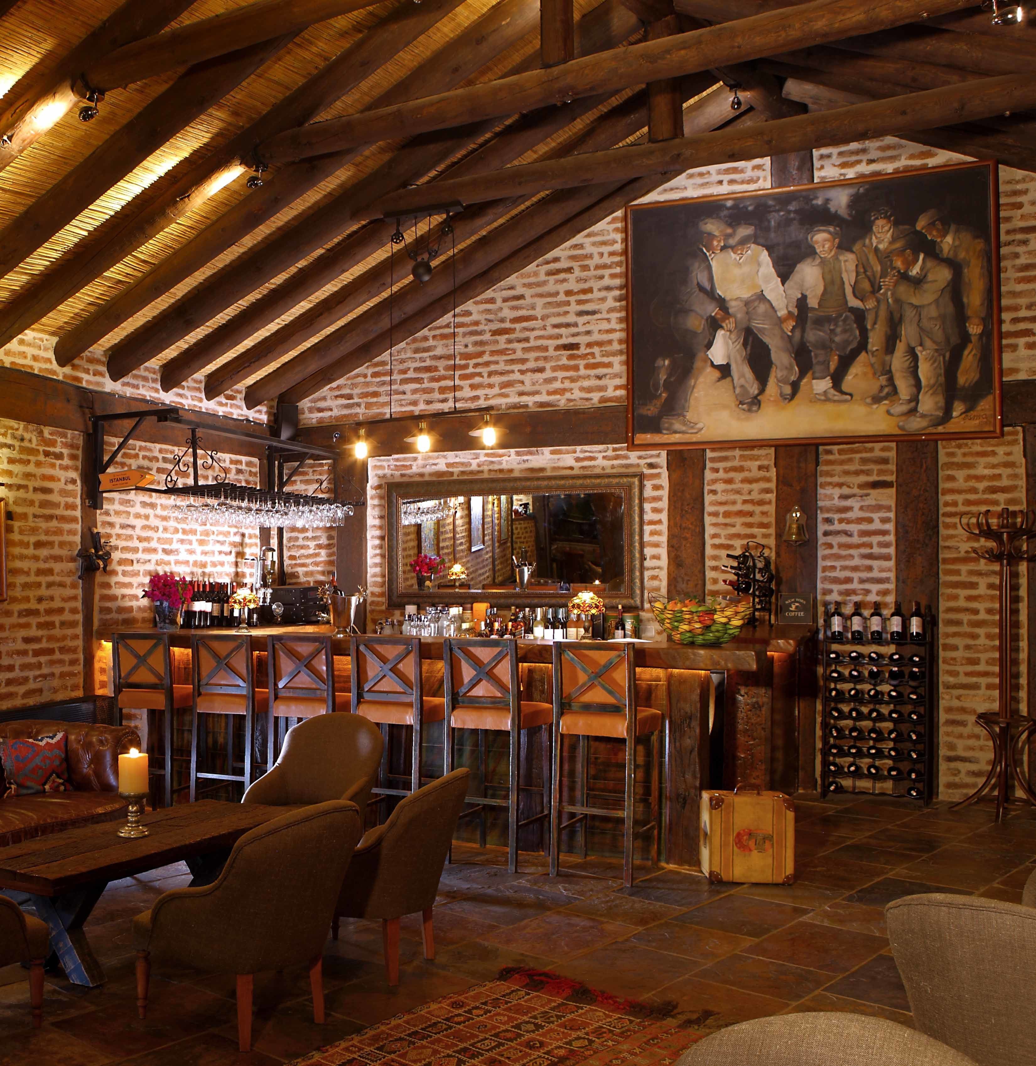 A rustic bar area with wooden beams, brick walls, and a large painting. There are bar stools, a table with chairs, and a wine rack.