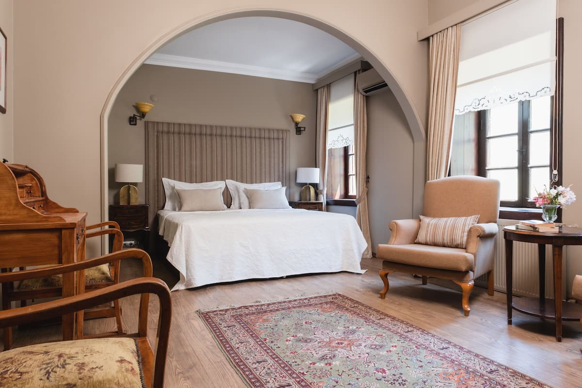 A hotel room with a bed, a wooden desk, a chair, a small table with a vase, and a large rug.