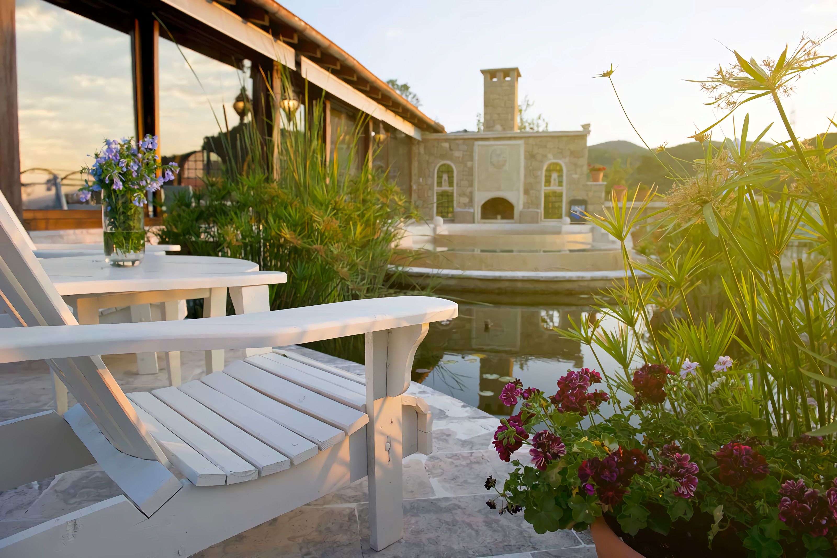 A patio area with white wooden chairs and a table, surrounded by potted plants and a stone building with arched windows in the background.