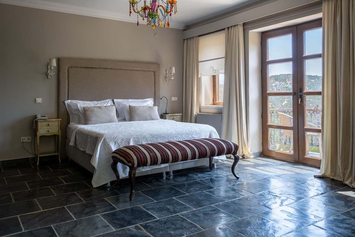 A spacious bedroom with a large bed, a bench, and a view of a hillside through a large window.