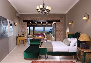 Quarto com uma cama grande, poltronas estofadas em verde, uma escrivaninha de madeira e vista através de uma porta de vidro.