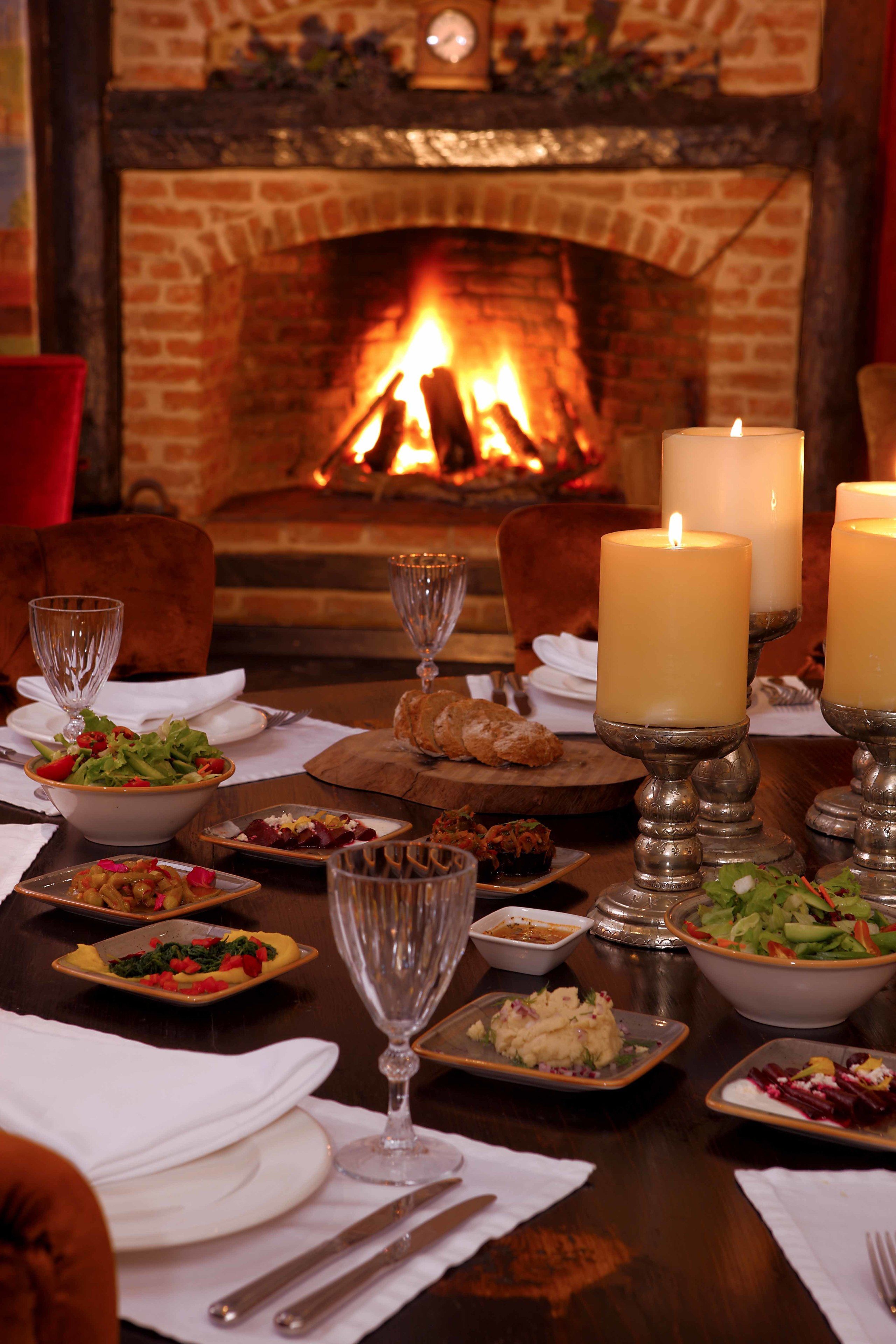 A dining table set for a meal with candles, plates of food, and a fireplace in the background.