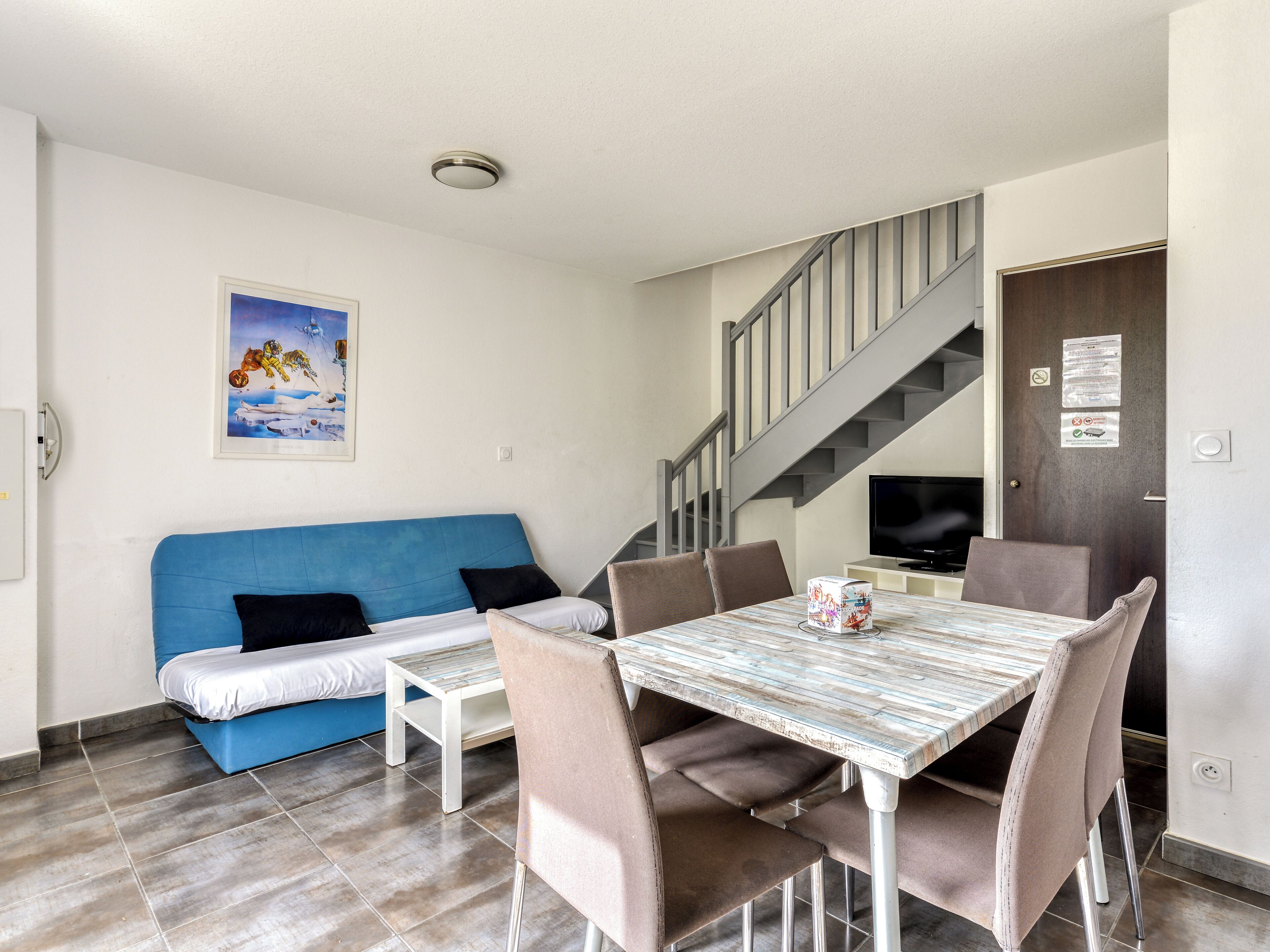 Una habitación con un sofá azul, una mesa de comedor con sillas, un televisor y una escalera.