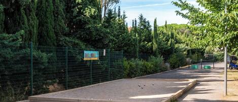Un'area sterrata con una piattaforma in legno, circondata da una recinzione verde e alberi rigogliosi.