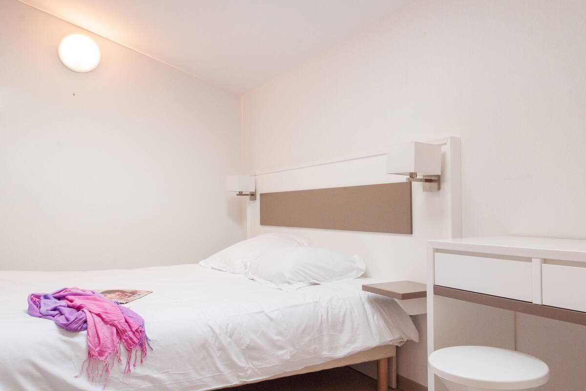Ein schlicht eingerichtetes Schlafzimmer mit Bett, Nachttisch und einem kleinen weißen Hocker.
