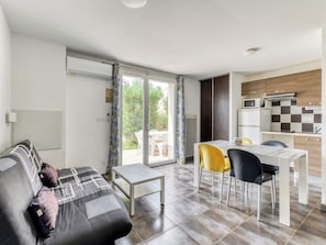 A modern living room with a sofa, a dining table with chairs, a kitchen area with a microwave and refrigerator, and a sliding glass door leading to an outdoor area.