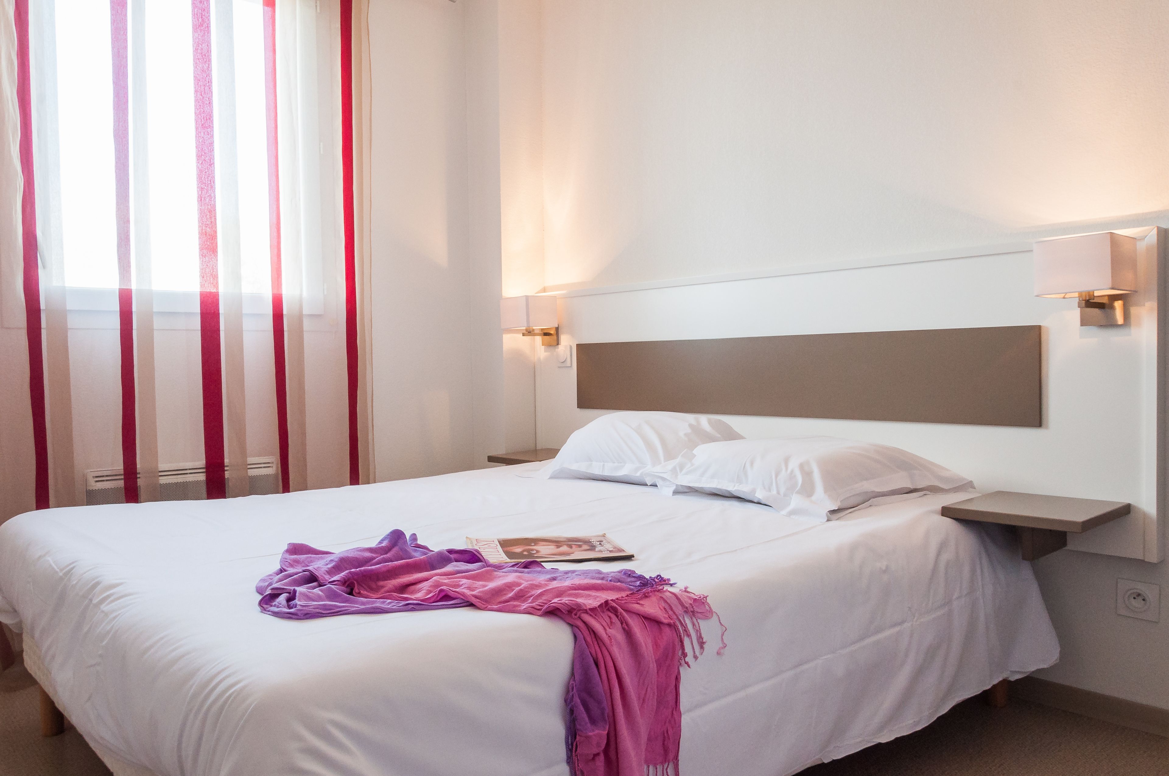 Una habitación de hotel con una cama grande, ropa de cama blanca, cabecera, lámparas de noche y una ventana con cortinas.