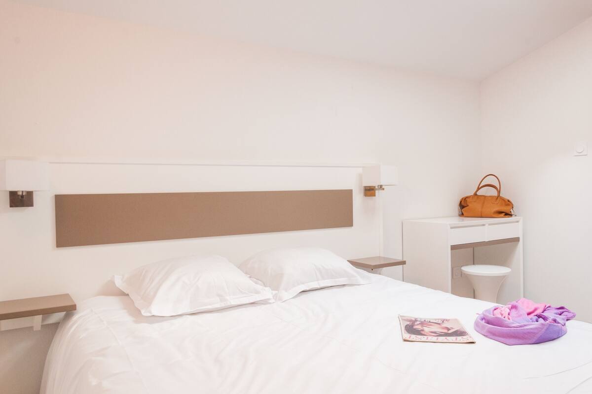 Ein ordentlich eingerichtetes Schlafzimmer mit Bett, Nachttisch, kleiner Schreibtisch und einem violetten Sitzsack.