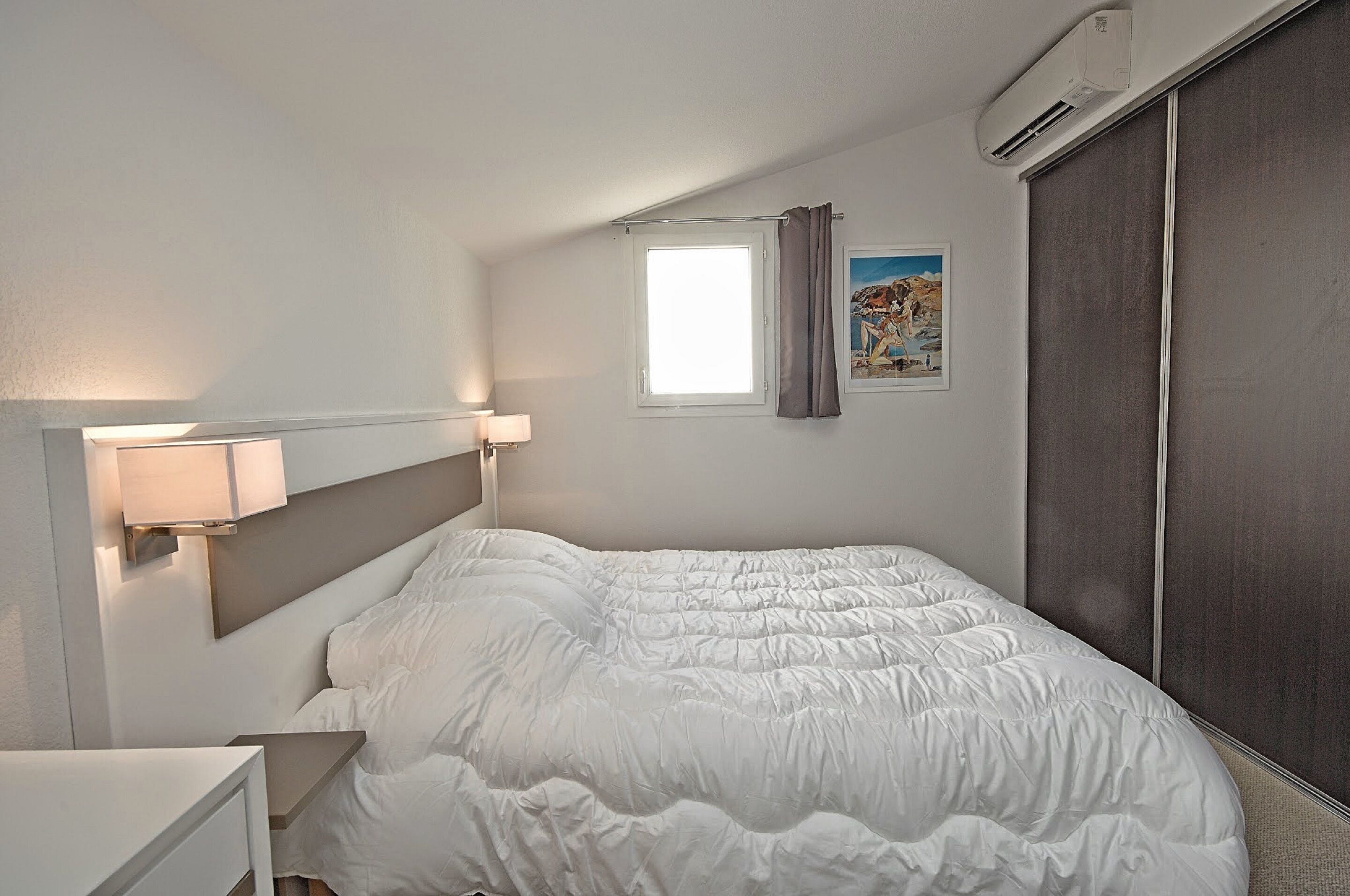 A neatly made bed with a white duvet, a bedside lamp, and a window with a curtain.