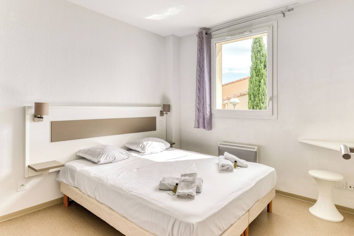 Ein modernes Schlafzimmer mit einem großen Bett, einem Nachttisch, einem Fenster mit Vorhängen, einer Heizung und einem kleinen weißen Hocker.
