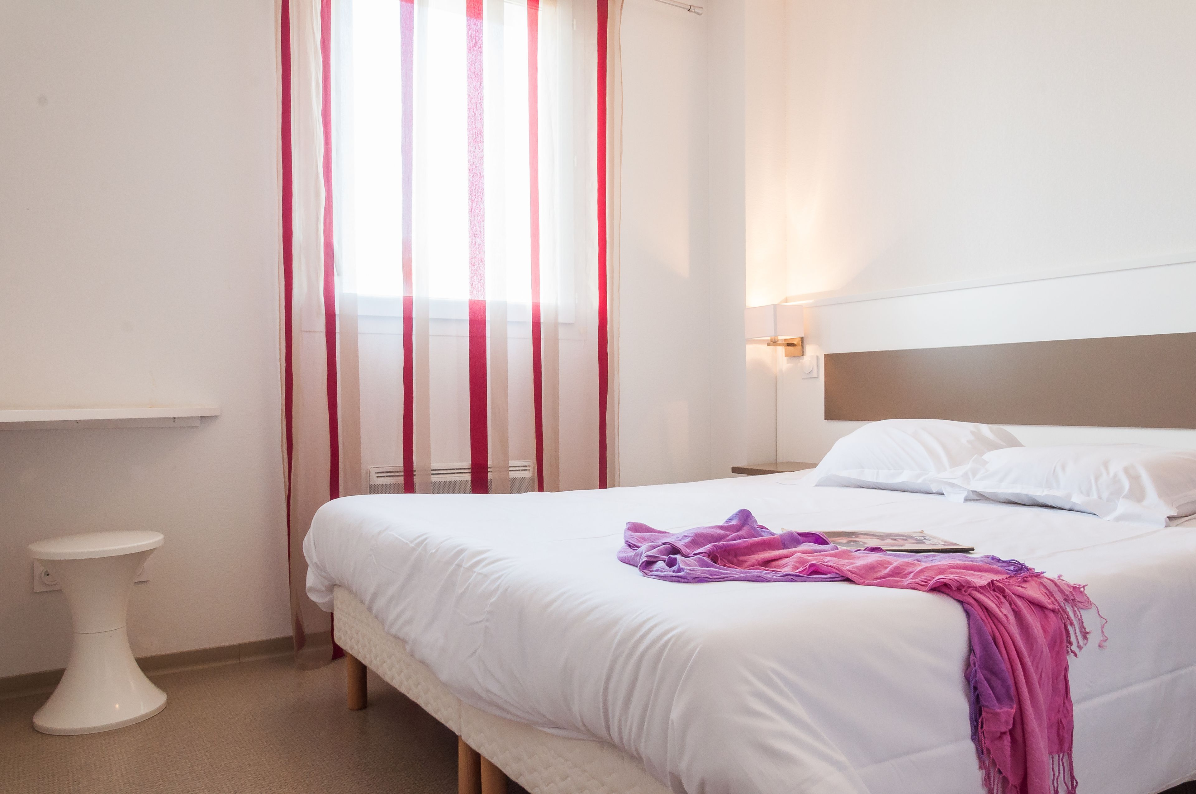 A neatly made bed with a white headboard, a bedside table, and a window with red curtains.