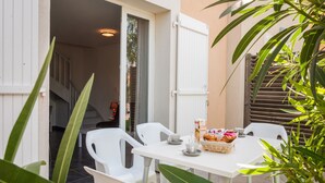 Une terrasse avec une table dressée pour deux personnes, des chaises blanches, un panier de pain et une boîte de céréales.