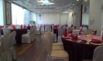 Un salón de banquetes con filas de mesas dispuestas para un evento formal, con sillas blancas adornadas con cintas doradas, servilletas rojas y una zona de escenario central.