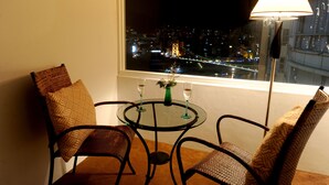 Een kamer met een glazen tafel, twee rieten stoelen, een vloerlamp en uitzicht op een stadsbeeld in de nacht.