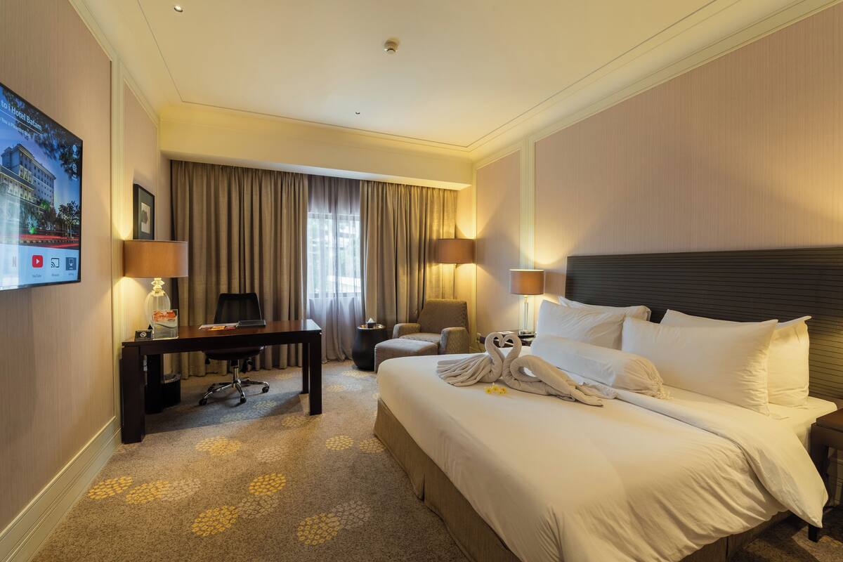 A hotel room with a bed, two bedside lamps, a desk with a computer, a chair, and a large window with curtains.