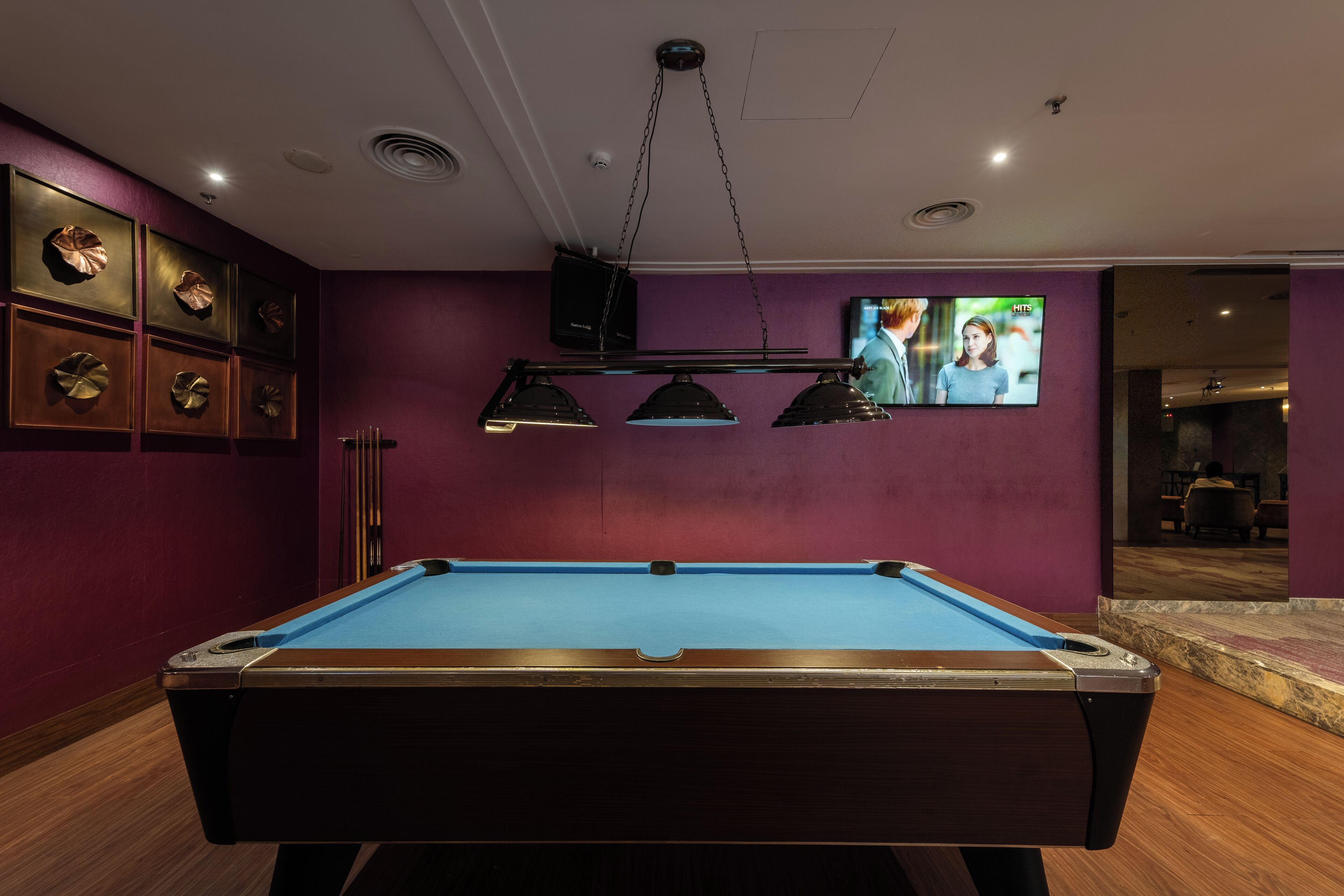Uma mesa de bilhar em um espaço com paredes roxas, um projetor fixado no teto e uma tela de televisão.