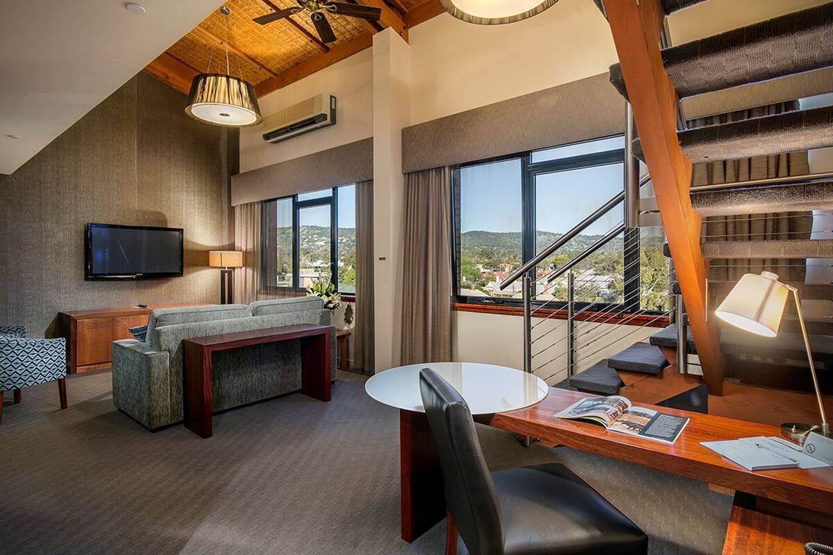 A modern hotel room with a large window, a flat-screen TV, a sofa, a dining table, and a staircase.