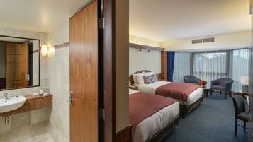 A hotel room with a wooden door, a bathroom with a sink, two beds with red bedding, a chair, and a window with curtains.