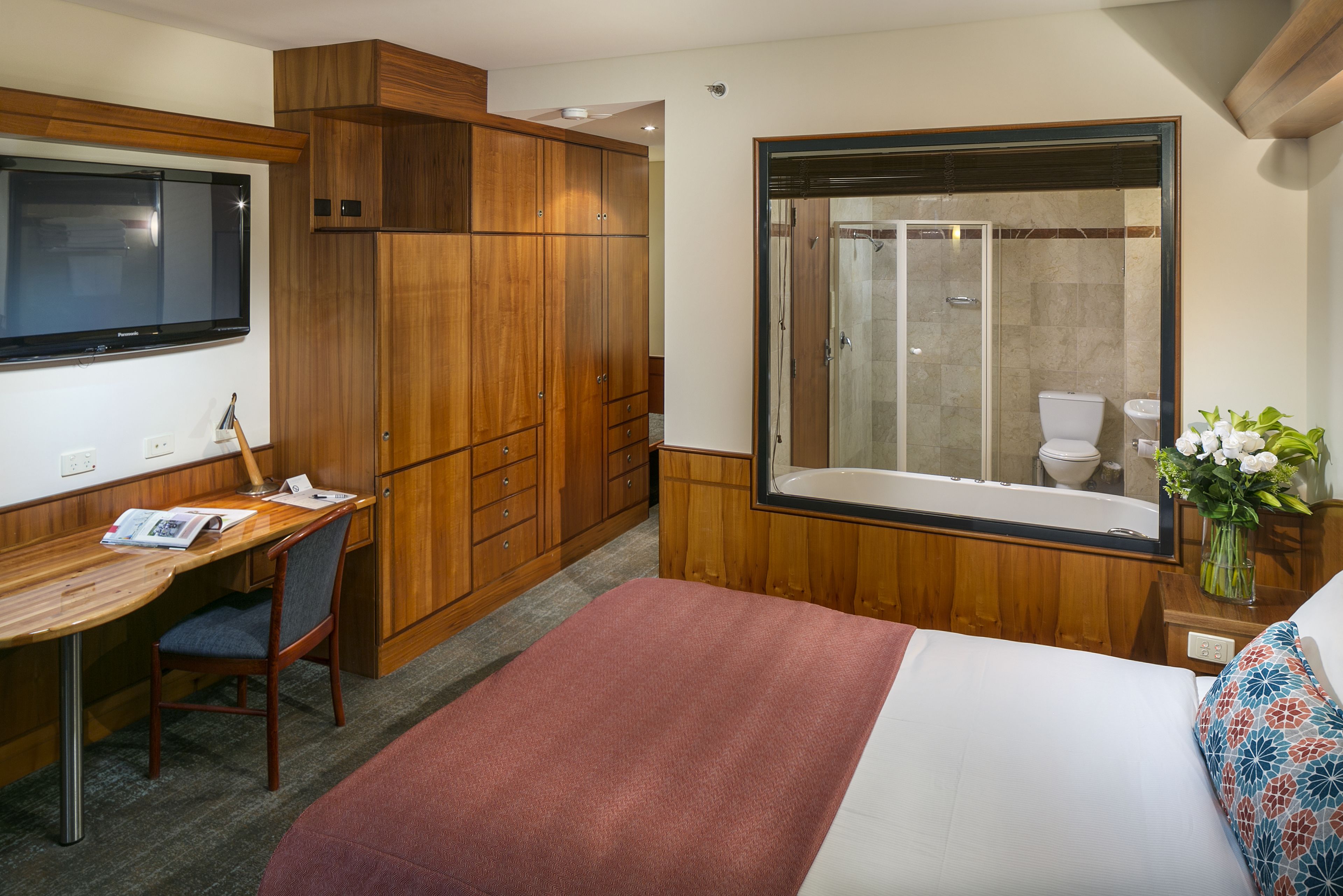 A hotel room with a wooden desk, a bed, a shower, and a large window.