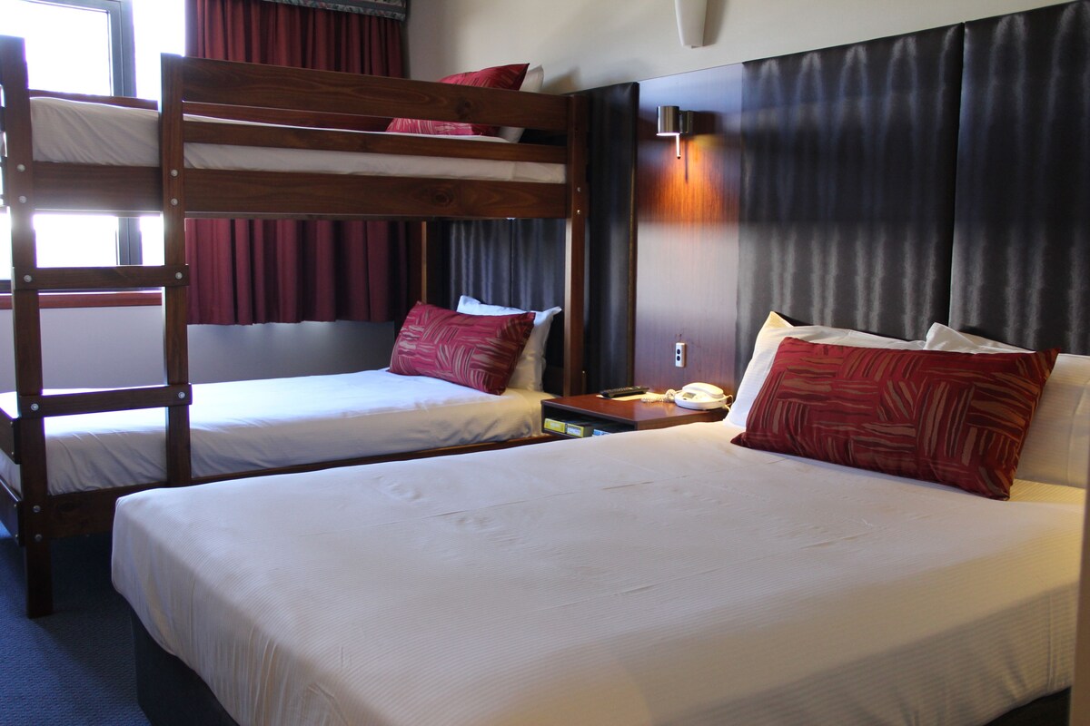 A hotel room with a bunk bed, two single beds, a bedside table with a lamp, and a window with curtains.
