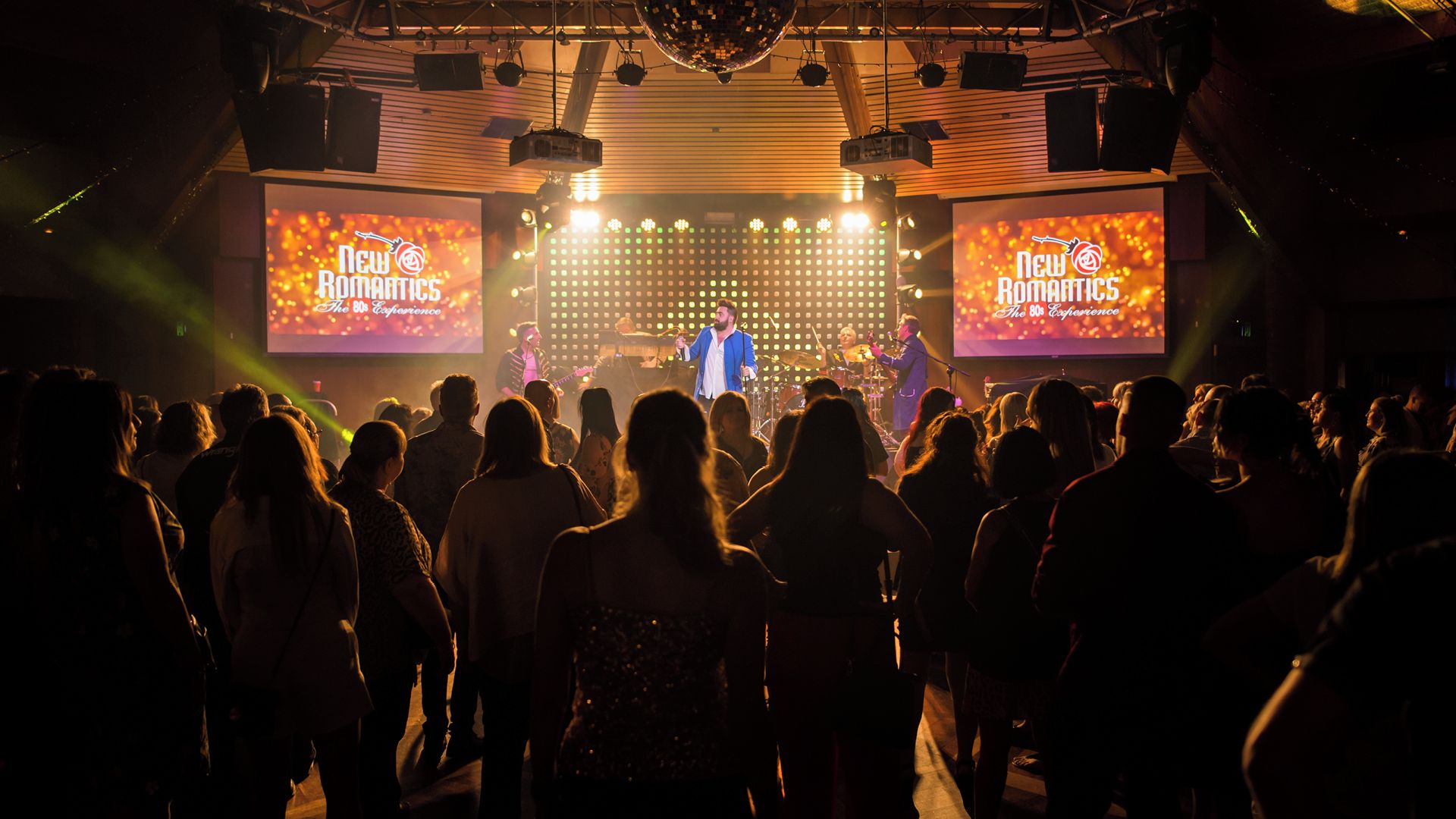 A concert with a stage, audience, and large screens displaying 'New Romantics The 80s Experience'.