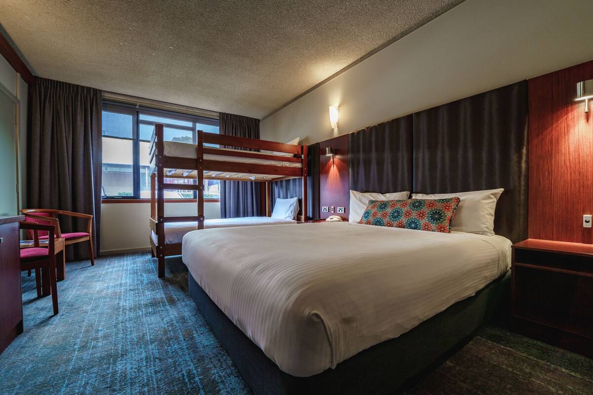 A hotel room with a bunk bed, a desk with a chair, a bedside table, and a window with curtains.