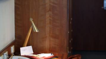A wooden desk with a lamp, papers, a telephone, and a chair in a room with wooden walls and a door in the background.