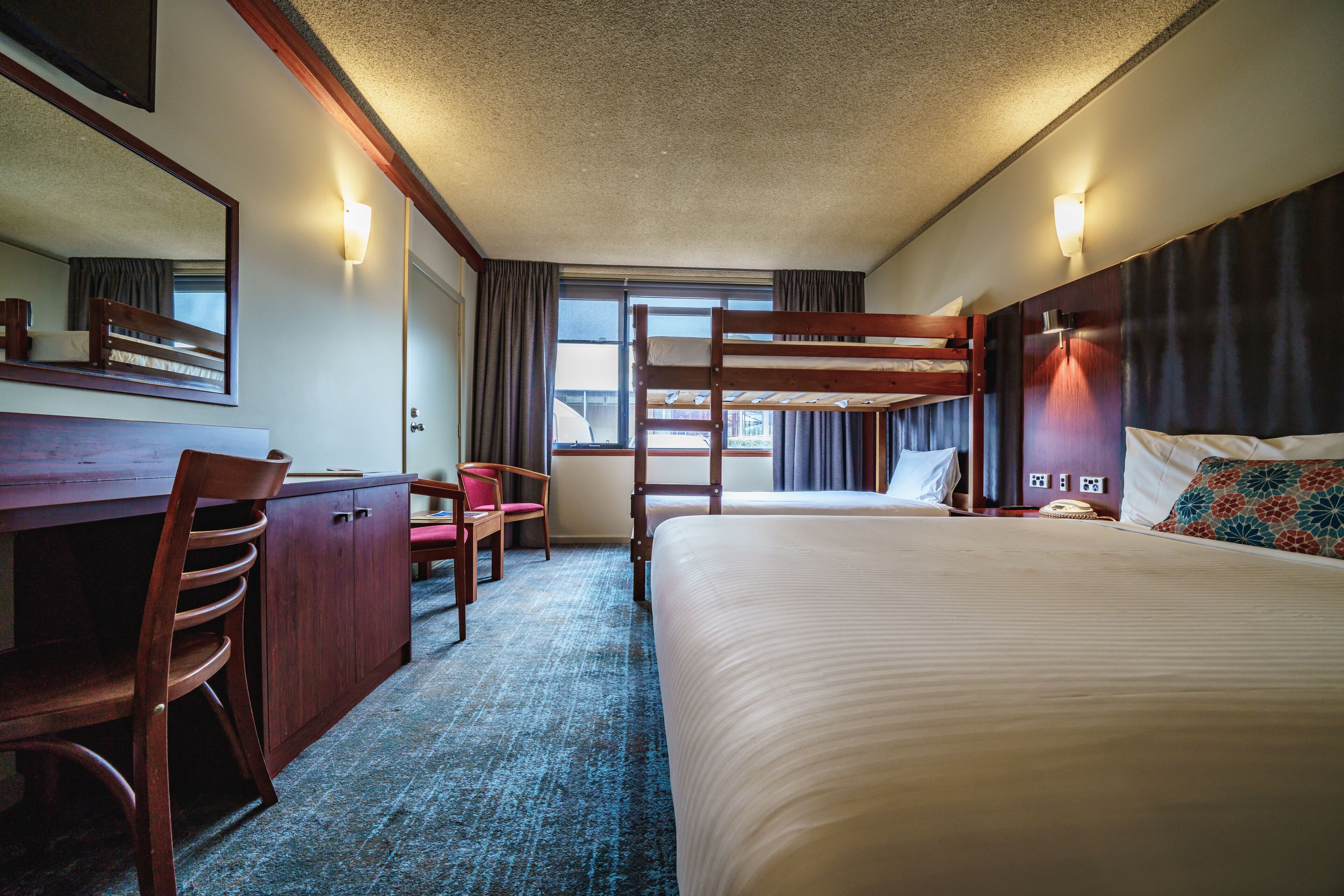 A hotel room with bunk beds, a desk with a chair, a mirror, and a window with curtains.