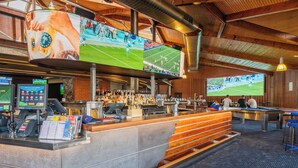Zona de bar amb diverses pantalles que mostren partits esportius, taula de bitlles i barra amb un menú i terminals de pagament.