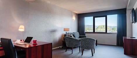 Habitación de hotel moderna con un escritorio de madera, una computadora portátil, una taza roja, dos sillas estampadas, un sofá y un ventanal con vistas al exterior.