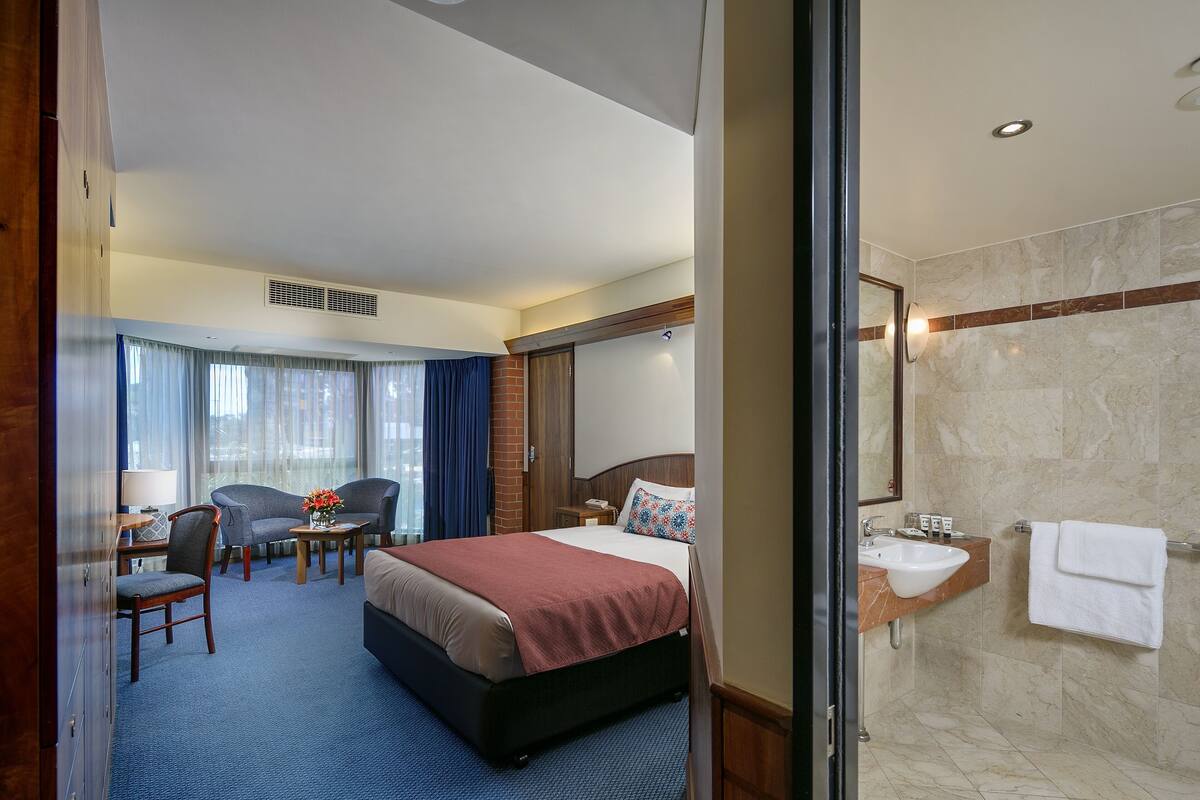 A hotel room with a bed, a desk with a chair, a small table with a vase, and a bathroom with a sink and mirror.