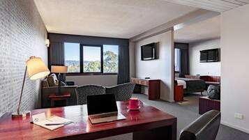 A hotel room with a wooden desk, a laptop, a red mug, a bed, a TV, and a view of the outdoors.