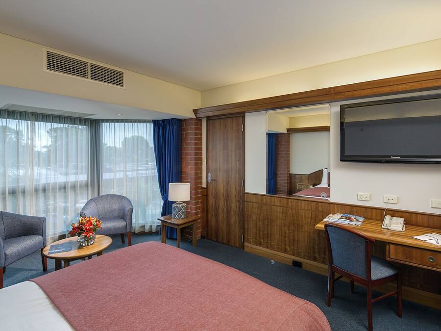 Quarto de hotel com cama, duas poltronas, escrivaninha com televisão, espelho e janela com cortinas.