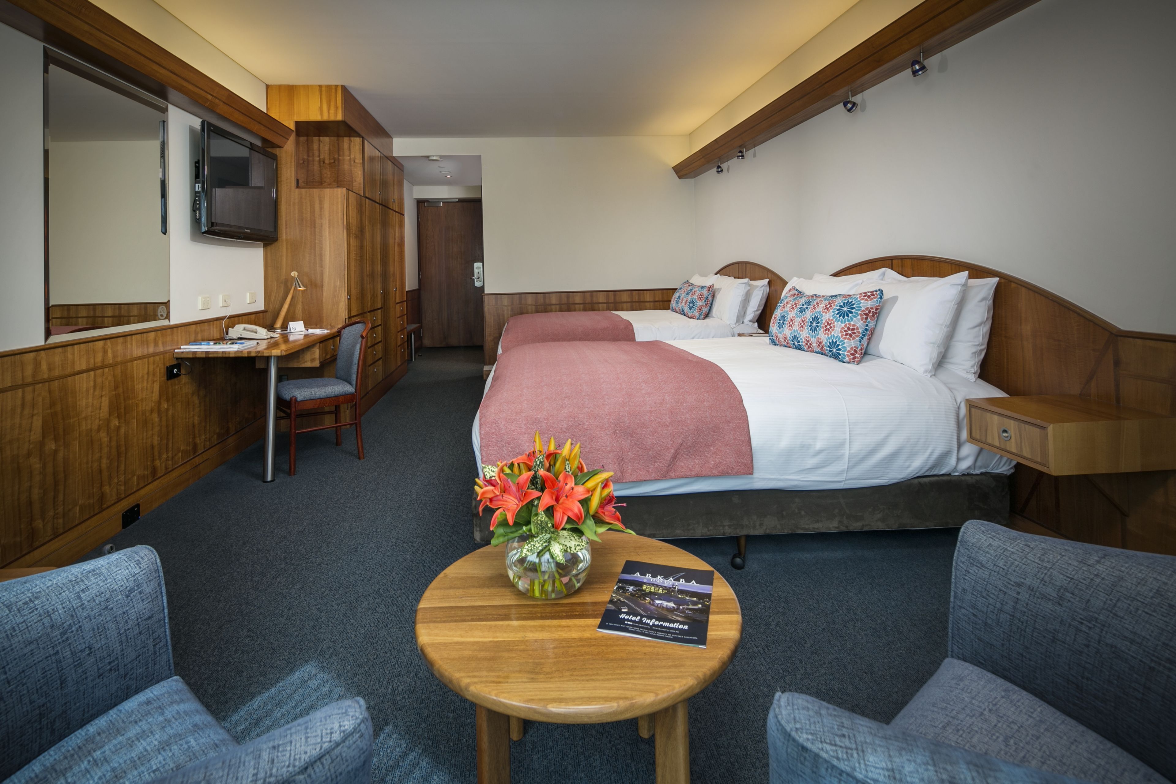 A hotel room with a bed, a desk, a chair, a small table with a vase of flowers, and a television mounted on the wall.