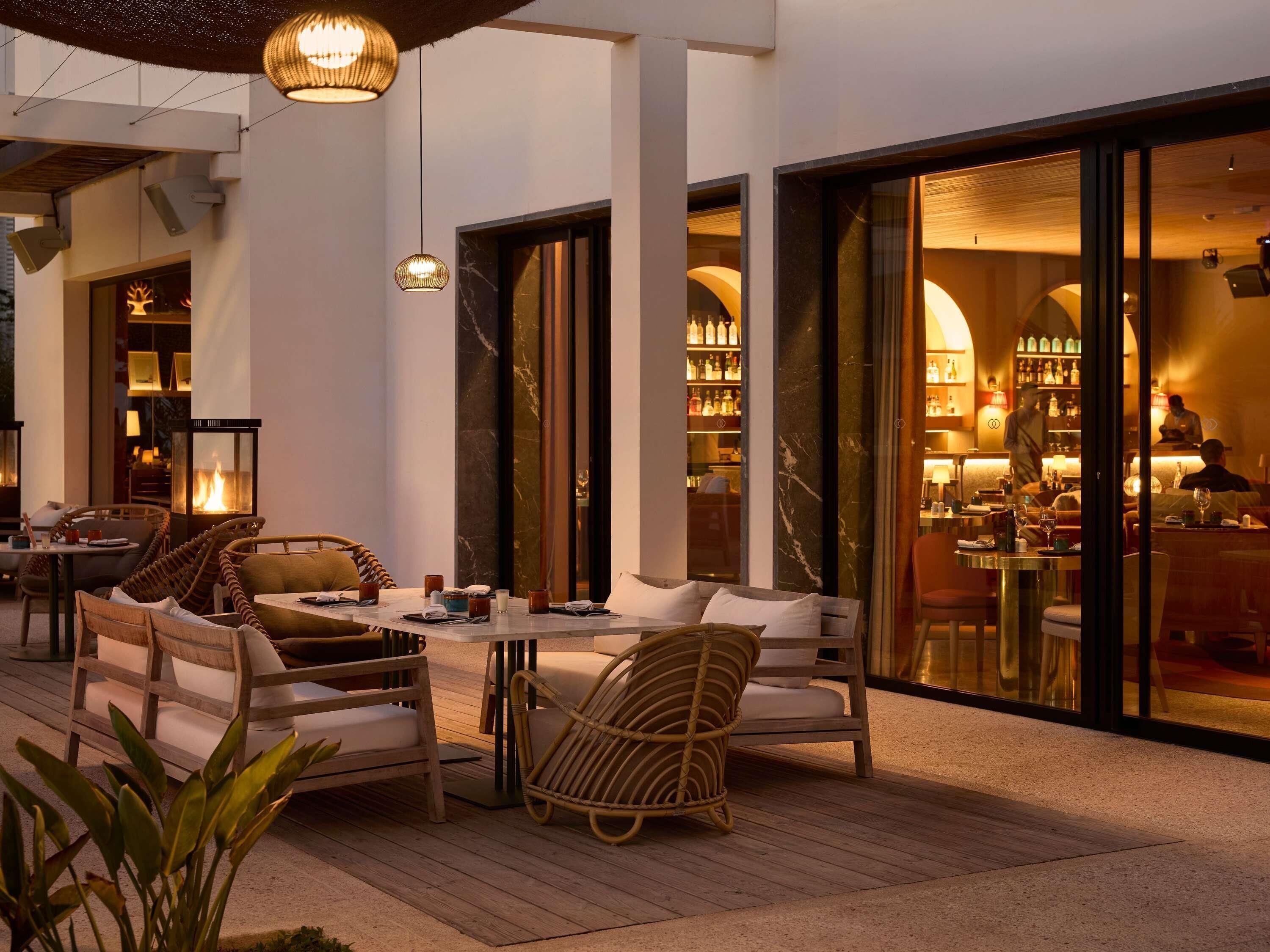 An outdoor seating area with wicker furniture, a fire feature, and a bar visible through glass doors.