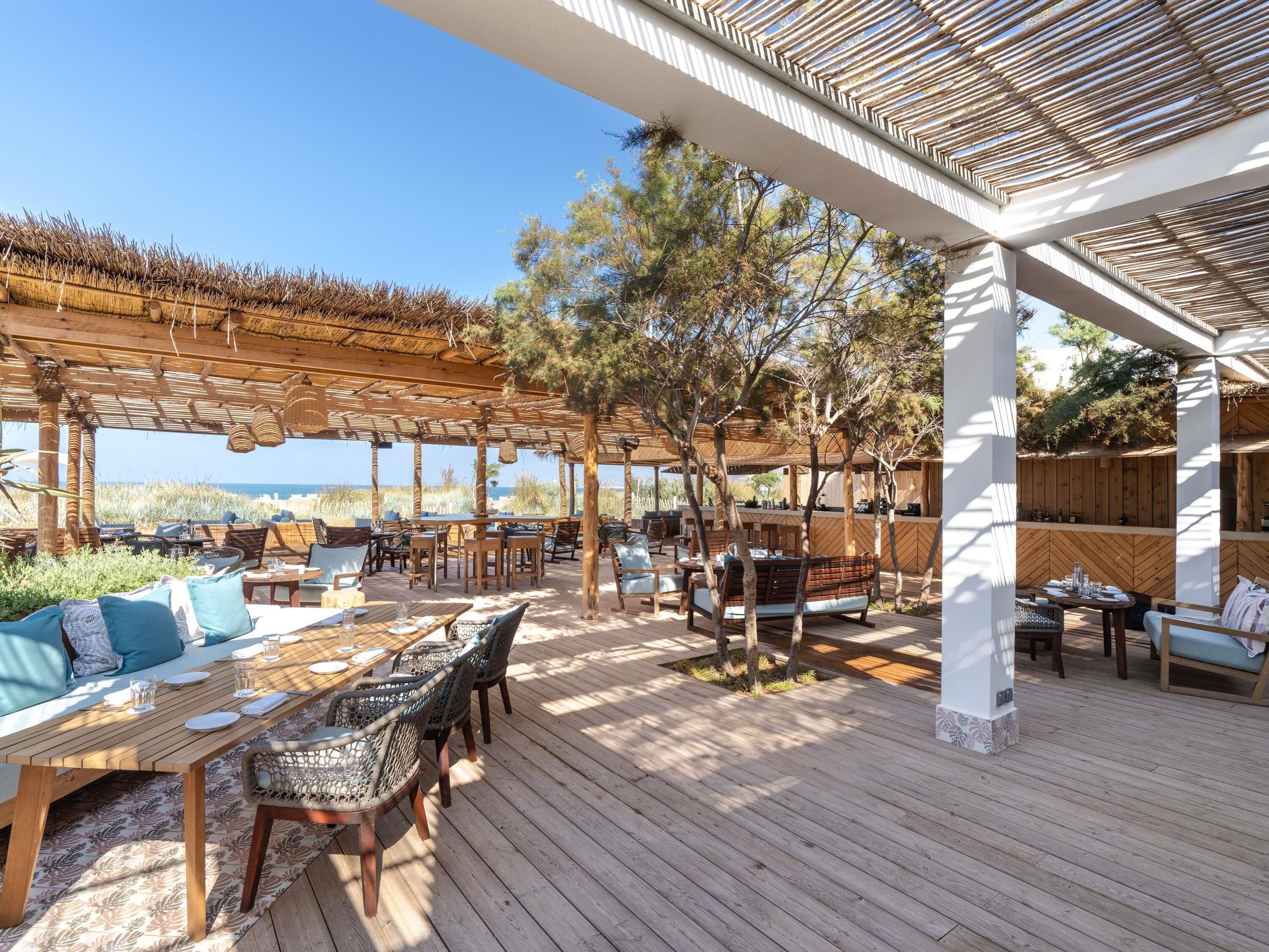 Un patio al aire libre con mobiliario de madera, techo de paja y vista al mar.