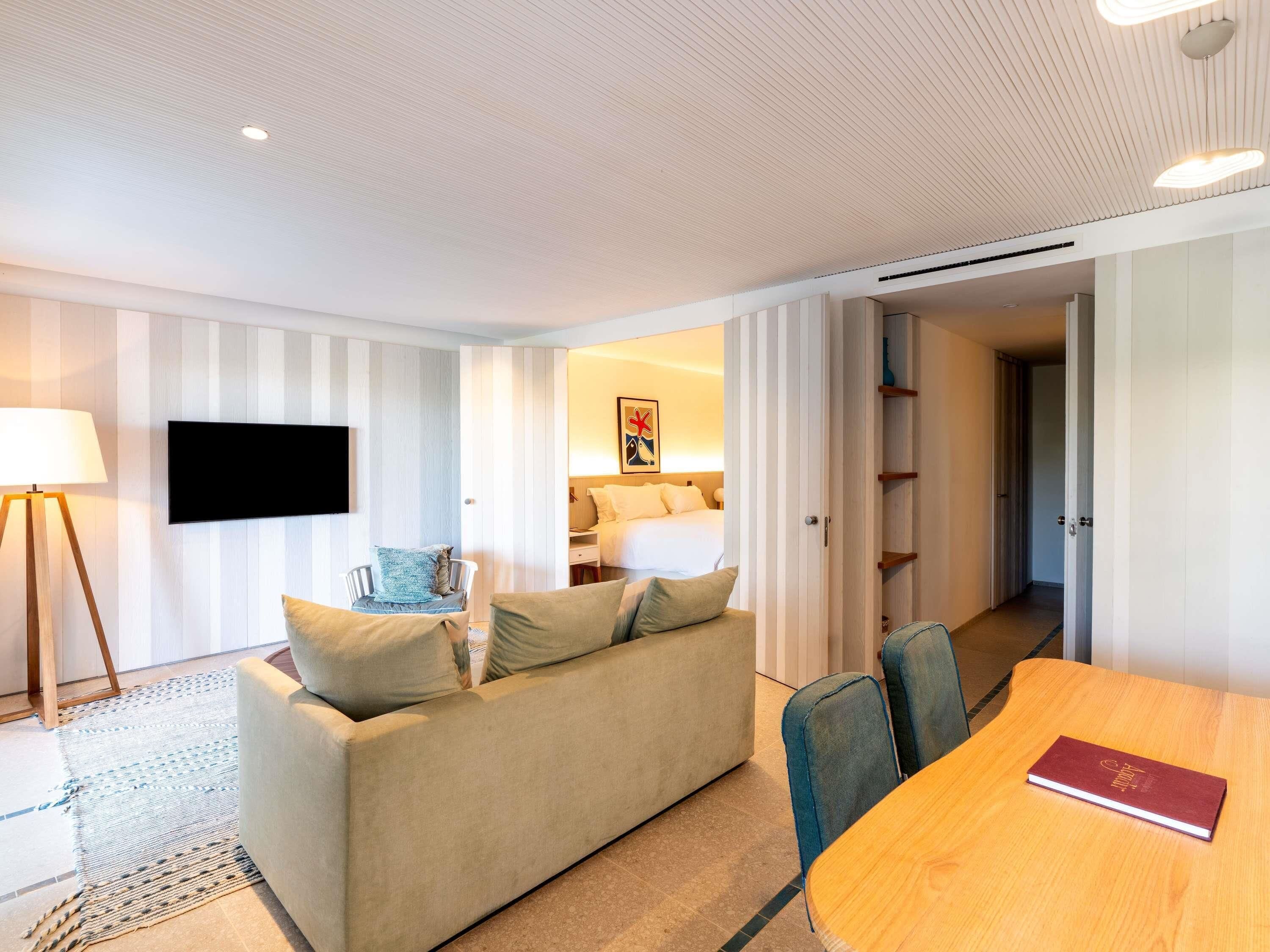 A modern hotel room with a sofa, a TV, a dining table, and a bookshelf.
